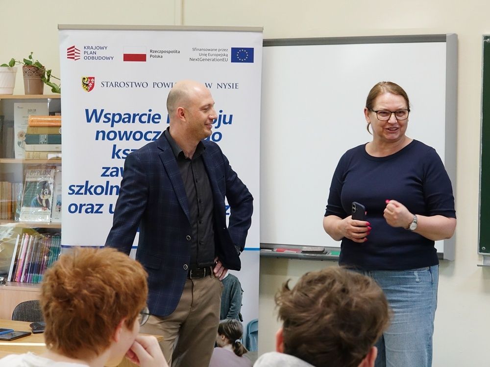 Zdjęcie przedstawia dwoje prelegentów – mężczyznę w marynarce i kobietę w ciemnej bluzce – przemawiających do grupy uczniów w klasie. Prelegenci stoją obok baneru promocyjnego 