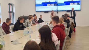 Grupa młodych ludzi siedzi wokół długiego stołu konferencyjnego, słuchając kobiety stojącej z przodu pokoju. W pobliżu stoi mężczyzna. Dwa duże ekrany wyświetlają stronę internetową. Na stole leżą butelki wody, notatniki i długopisy. Pokój jest jasny z dużymi oknami.