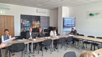 Uczestnicy spotkania siedzą przy złączonych stołach w sali z tablicą informacyjną o regionie Opolszczyzny. Jedna osoba stoi z boku i czyta dokumenty, podczas gdy reszta grupy jest skupiona na pracy indywidualnej. Wnętrze jest nowoczesne, wyposażone w klimatyzację i oznakowane wyjście ewakuacyjne.