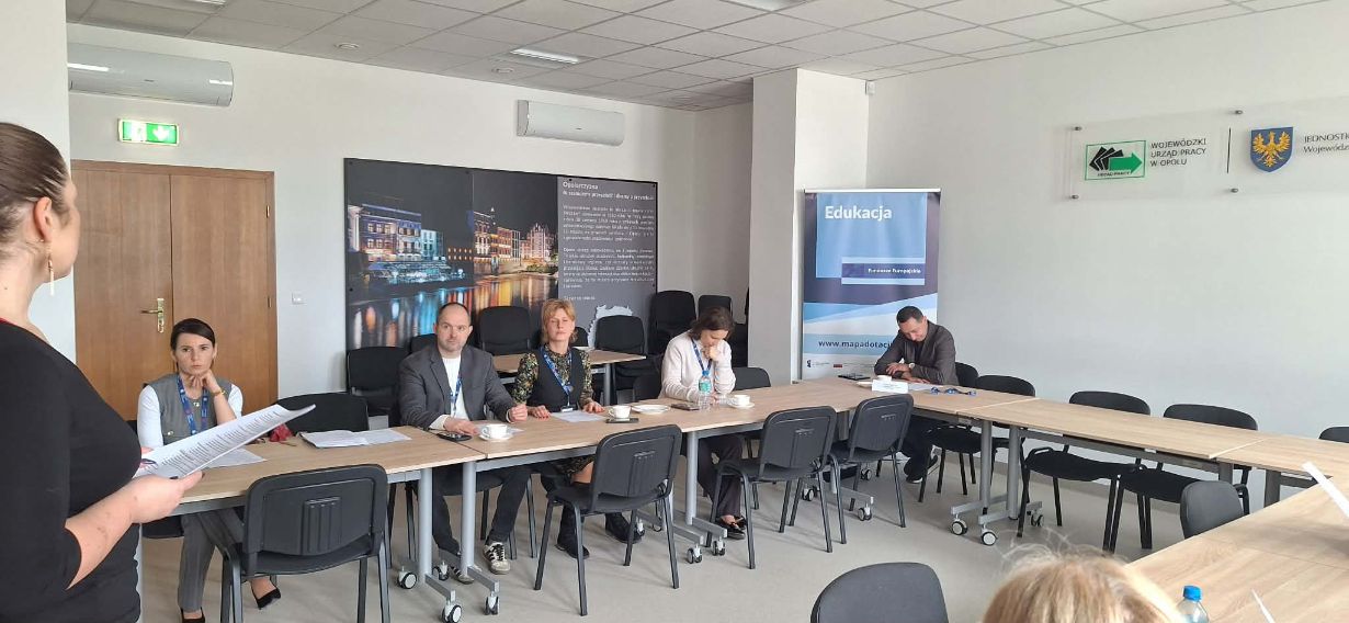 Uczestnicy spotkania siedzą przy złączonych stołach w sali z tablicą informacyjną o regionie Opolszczyzny. Jedna osoba stoi z boku i czyta dokumenty, podczas gdy reszta grupy jest skupiona na pracy indywidualnej. Wnętrze jest nowoczesne, wyposażone w klimatyzację i oznakowane wyjście ewakuacyjne.