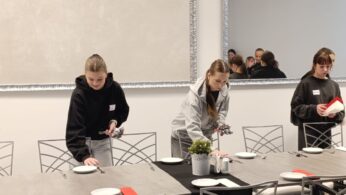 Trzy dziewczyny w bluzach z imiennymi plakietkami przygotowują nakrycia na długim stole w eleganckiej sali. Starannie rozkładają talerze, sztućce i serwetki, dbając o estetykę stanowisk. W tle widać duże lustro odbijające fragmenty nowoczesnego wnętrza.