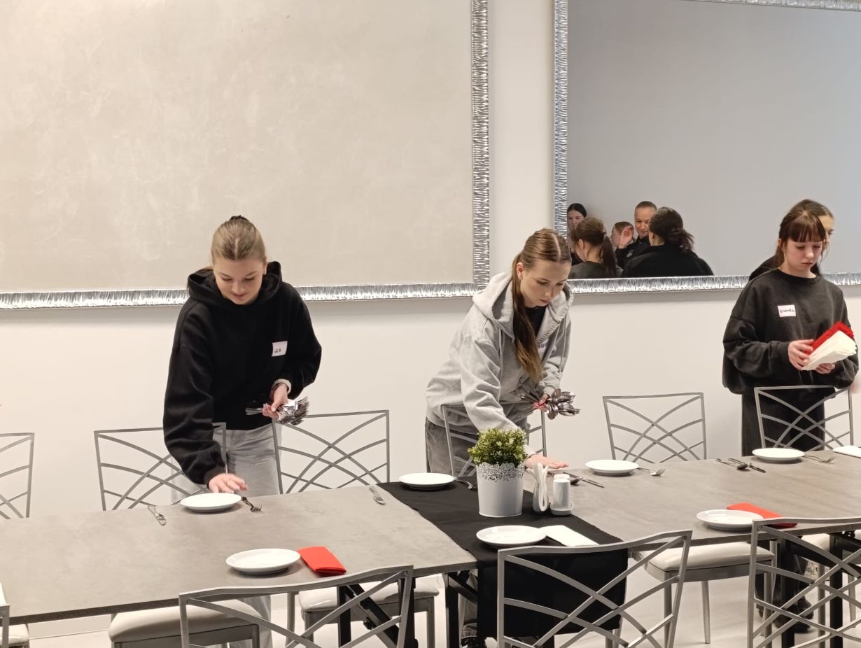 Trzy dziewczyny w bluzach z imiennymi plakietkami przygotowują nakrycia na długim stole w eleganckiej sali. Starannie rozkładają talerze, sztućce i serwetki, dbając o estetykę stanowisk. W tle widać duże lustro odbijające fragmenty nowoczesnego wnętrza.
