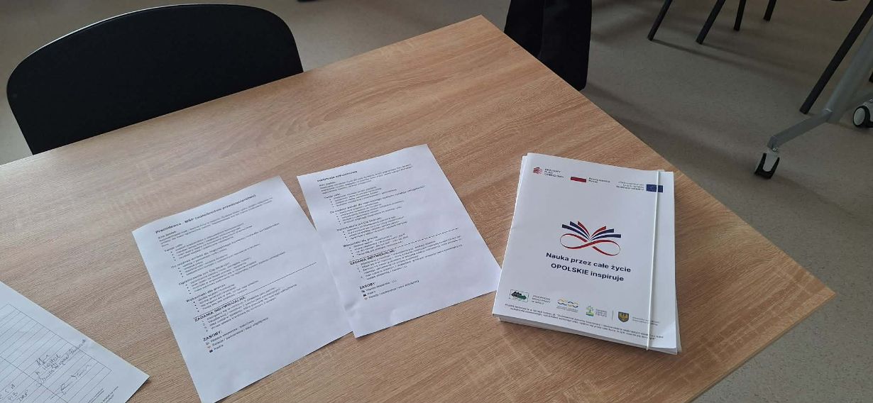 Zbliżenie na dokumenty szkoleniowe leżące na drewnianym stole, w tym stos broszur z logo „Nauka przez całe życie”. Na kartkach widoczne są nagłówki dotyczące ról pracodawcy i instytucji szkoleniowej w systemie rozwoju umiejętności. Materiały są opatrzone logotypami Funduszy Europejskich oraz Rzeczpospolitej Polskiej.