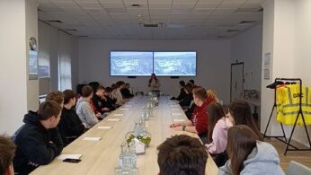 Grupa ludzi siedzi wokół długiego stołu konferencyjnego w jasnej sali konferencyjnej, słuchając prezentera z przodu. Dwa ekrany wyświetlają slajdy za prezenterem. Na stole leżą butelki z wodą, szklanki i notatniki. Kamizelki odblaskowe wiszą na stojaku.