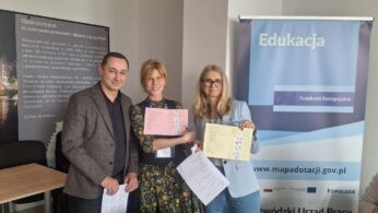 Dwie kobiety i mężczyzna pozują do zdjęcia, trzymając w rękach kolorowe arkusze dokumentów i długopisy. Stoją na tle tablicy o Opolszczyźnie oraz niebieskiego baneru z napisem „Edukacja – Fundusze Europejskie”. Wszyscy są uśmiechnięci i ubrani w stroje o charakterze biznesowym.