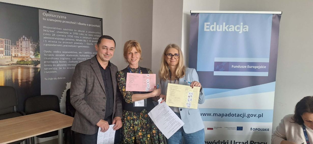 Dwie kobiety i mężczyzna pozują do zdjęcia, trzymając w rękach kolorowe arkusze dokumentów i długopisy. Stoją na tle tablicy o Opolszczyźnie oraz niebieskiego baneru z napisem „Edukacja – Fundusze Europejskie”. Wszyscy są uśmiechnięci i ubrani w stroje o charakterze biznesowym.