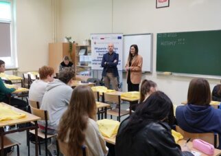 Ujęcie z perspektywy klasy, pokazujące prelegenta w marynarce i prelegentkę w beżowym żakiecie przemawiających do siedzących uczniów. W tle znajduje się baner promocyjny oraz zielona tablica z napisem "23.03.2026" i "WITAMY". Uczniowie w ławkach słuchają wystąpień, niektórzy mają żółte torby.