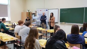 Ujęcie z perspektywy klasy, pokazujące prelegenta w marynarce i prelegentkę w beżowym żakiecie przemawiających do siedzących uczniów. W tle znajduje się baner promocyjny oraz zielona tablica z napisem 