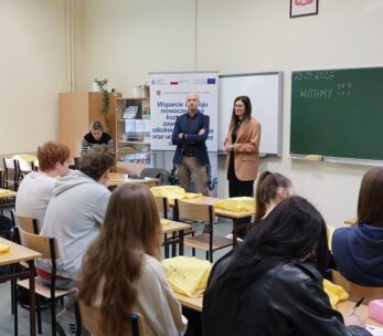 Ujęcie z perspektywy klasy, pokazujące prelegenta w marynarce i prelegentkę w beżowym żakiecie przemawiających do siedzących uczniów. W tle znajduje się baner promocyjny oraz zielona tablica z napisem "23.03.2026" i "WITAMY". Uczniowie w ławkach słuchają wystąpień, niektórzy mają żółte torby.