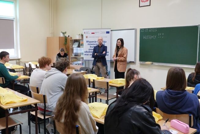 Ujęcie z perspektywy klasy, pokazujące prelegenta w marynarce i prelegentkę w beżowym żakiecie przemawiających do siedzących uczniów. W tle znajduje się baner promocyjny oraz zielona tablica z napisem "23.03.2026" i "WITAMY". Uczniowie w ławkach słuchają wystąpień, niektórzy mają żółte torby.