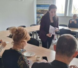 Perspektywa zza pleców jednego z uczestników na grupę osób pracującą przy wspólnym stole. Prowadząca spotkanie stoi pośrodku i przekazuje dokumenty siedzącej kobiecie. W tle, przy oknie, pozostali uczestnicy analizują otrzymane materiały w świetle dziennym.