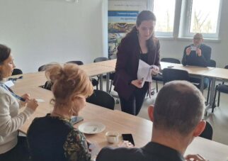 Perspektywa zza pleców jednego z uczestników na grupę osób pracującą przy wspólnym stole. Prowadząca spotkanie stoi pośrodku i przekazuje dokumenty siedzącej kobiecie. W tle, przy oknie, pozostali uczestnicy analizują otrzymane materiały w świetle dziennym.