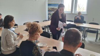 Perspektywa zza pleców jednego z uczestników na grupę osób pracującą przy wspólnym stole. Prowadząca spotkanie stoi pośrodku i przekazuje dokumenty siedzącej kobiecie. W tle, przy oknie, pozostali uczestnicy analizują otrzymane materiały w świetle dziennym.