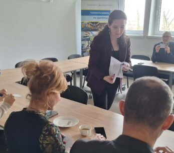 Perspektywa zza pleców jednego z uczestników na grupę osób pracującą przy wspólnym stole. Prowadząca spotkanie stoi pośrodku i przekazuje dokumenty siedzącej kobiecie. W tle, przy oknie, pozostali uczestnicy analizują otrzymane materiały w świetle dziennym.