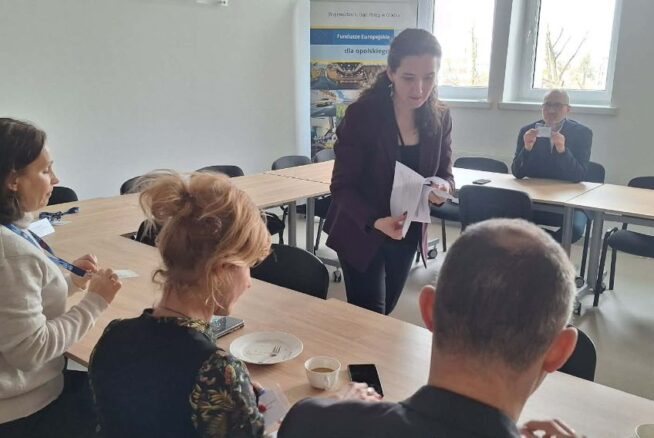 Perspektywa zza pleców jednego z uczestników na grupę osób pracującą przy wspólnym stole. Prowadząca spotkanie stoi pośrodku i przekazuje dokumenty siedzącej kobiecie. W tle, przy oknie, pozostali uczestnicy analizują otrzymane materiały w świetle dziennym.