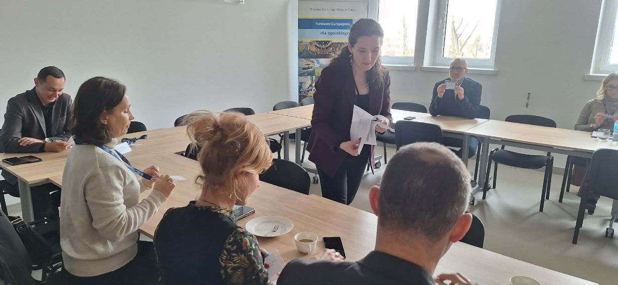 Perspektywa zza pleców jednego z uczestników na grupę osób pracującą przy wspólnym stole. Prowadząca spotkanie stoi pośrodku i przekazuje dokumenty siedzącej kobiecie. W tle, przy oknie, pozostali uczestnicy analizują otrzymane materiały w świetle dziennym.