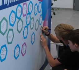 Szerokie ujęcie nowoczesnej hali, w której odbywa się Festiwal Kariery. Na pierwszym planie widać liczne grupy młodzieży siedzące na kolorowych pufach oraz stoiska wystawiennicze w głębi. Nad wejściem widnieje duży napis „WEJŚCIE NA TRYBUNY”, a cała przestrzeń jest jasno oświetlona.