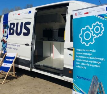 Biała furgonetka z napisem EDUBUS jest zaparkowana na zewnątrz w słoneczny dzień. Obok niego znajdują się dwa niebieskie leżaki i wysoki niebieski znak z białymi ikonami sprzętu i polskim tekstem. W tle widoczne są drzewa, budynki i niebieski namiot.