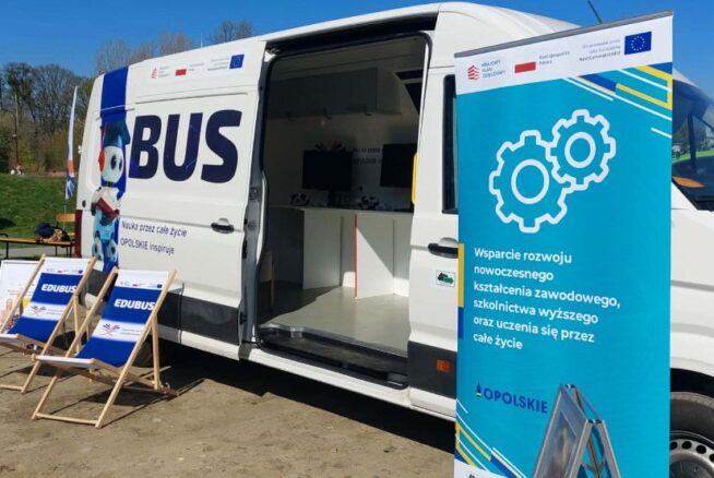 Biała furgonetka z napisem EDUBUS jest zaparkowana na zewnątrz w słoneczny dzień. Obok niego znajdują się dwa niebieskie leżaki i wysoki niebieski znak z białymi ikonami sprzętu i polskim tekstem. W tle widoczne są drzewa, budynki i niebieski namiot.