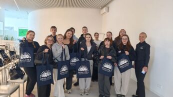 Grupa młodzieży pozuje do wspólnego zdjęcia w jasnym, nowoczesnym lobby z charakterystycznym ażurowym sufitem. Większość osób trzyma granatowe torby bawełniane z białym logo 