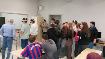 Grupa młodzieży szkolnej oraz dorosłych obserwuje dwóch uczniów korzystających z gogli VR w jasnej klasie. Uczniowie z goglami stoją przy stanowiskach z monitorami, a reszta grupy przygląda się ich działaniom z zainteresowaniem. W tle widoczny jest interaktywny ekran oraz biała tablica, a jeden z uczniów na pierwszym planie ma na sobie koszulkę piłkarską Bayernu Monachium.