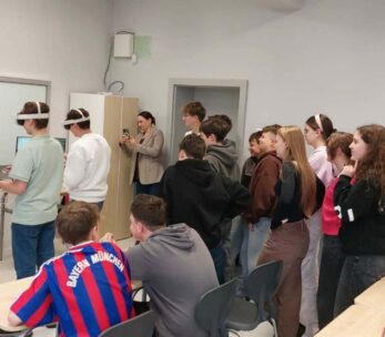 Grupa młodzieży szkolnej oraz dorosłych obserwuje dwóch uczniów korzystających z gogli VR w jasnej klasie. Uczniowie z goglami stoją przy stanowiskach z monitorami, a reszta grupy przygląda się ich działaniom z zainteresowaniem. W tle widoczny jest interaktywny ekran oraz biała tablica, a jeden z uczniów na pierwszym planie ma na sobie koszulkę piłkarską Bayernu Monachium.