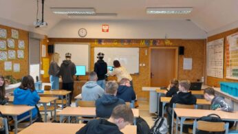 Szerokie ujęcie sali lekcyjnej, w której grupa uczniów uczestniczy w zajęciach z wykorzystaniem gogli VR. Na przedzie sali, pod napisem „TRENING CZYNI MISTRZA”, dwaj uczniowie testują technologię przy wsparciu opiekunów, podczas gdy reszta klasy siedzi w ławkach. Na ścianach wiszą pomoce naukowe, w tym układ okresowy pierwiastków oraz prace plastyczne. Pomieszczenie jest jasne, wyposażone w rzutnik podwieszony pod sufitem.