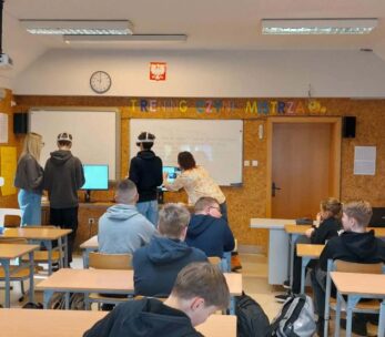 Szerokie ujęcie sali lekcyjnej, w której grupa uczniów uczestniczy w zajęciach z wykorzystaniem gogli VR. Na przedzie sali, pod napisem „TRENING CZYNI MISTRZA”, dwaj uczniowie testują technologię przy wsparciu opiekunów, podczas gdy reszta klasy siedzi w ławkach. Na ścianach wiszą pomoce naukowe, w tym układ okresowy pierwiastków oraz prace plastyczne. Pomieszczenie jest jasne, wyposażone w rzutnik podwieszony pod sufitem.