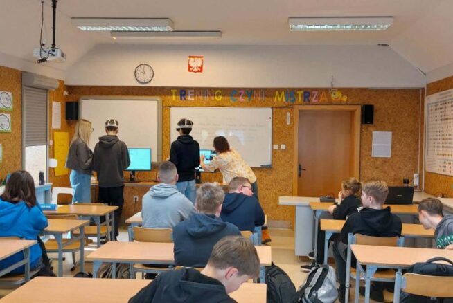 Szerokie ujęcie sali lekcyjnej, w której grupa uczniów uczestniczy w zajęciach z wykorzystaniem gogli VR. Na przedzie sali, pod napisem „TRENING CZYNI MISTRZA”, dwaj uczniowie testują technologię przy wsparciu opiekunów, podczas gdy reszta klasy siedzi w ławkach. Na ścianach wiszą pomoce naukowe, w tym układ okresowy pierwiastków oraz prace plastyczne. Pomieszczenie jest jasne, wyposażone w rzutnik podwieszony pod sufitem.