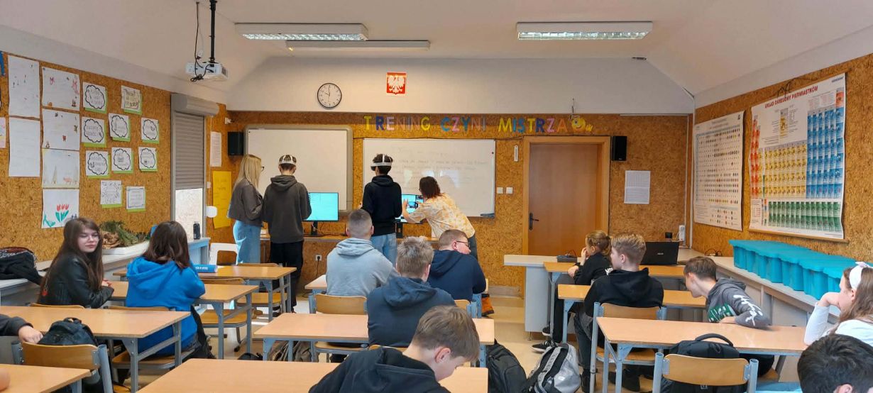 Szerokie ujęcie sali lekcyjnej, w której grupa uczniów uczestniczy w zajęciach z wykorzystaniem gogli VR. Na przedzie sali, pod napisem „TRENING CZYNI MISTRZA”, dwaj uczniowie testują technologię przy wsparciu opiekunów, podczas gdy reszta klasy siedzi w ławkach. Na ścianach wiszą pomoce naukowe, w tym układ okresowy pierwiastków oraz prace plastyczne. Pomieszczenie jest jasne, wyposażone w rzutnik podwieszony pod sufitem.