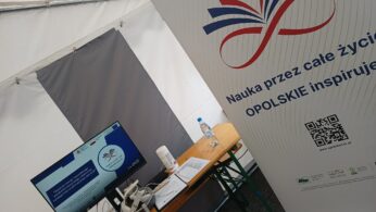 Stół wewnątrz namiotu mieści monitor komputerowy, zestaw VR, butelkę wody i inne przedmioty. Obok stoi wysoki baner z czerwono-niebieskim symbolem nieskończoności i polskim tekstem promującym uczenie się przez całe życie na Opolszczyźnie.