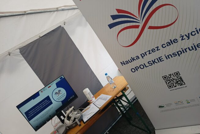 Stół wewnątrz namiotu mieści monitor komputerowy, zestaw VR, butelkę wody i inne przedmioty. Obok stoi wysoki baner z czerwono-niebieskim symbolem nieskończoności i polskim tekstem promującym uczenie się przez całe życie na Opolszczyźnie.