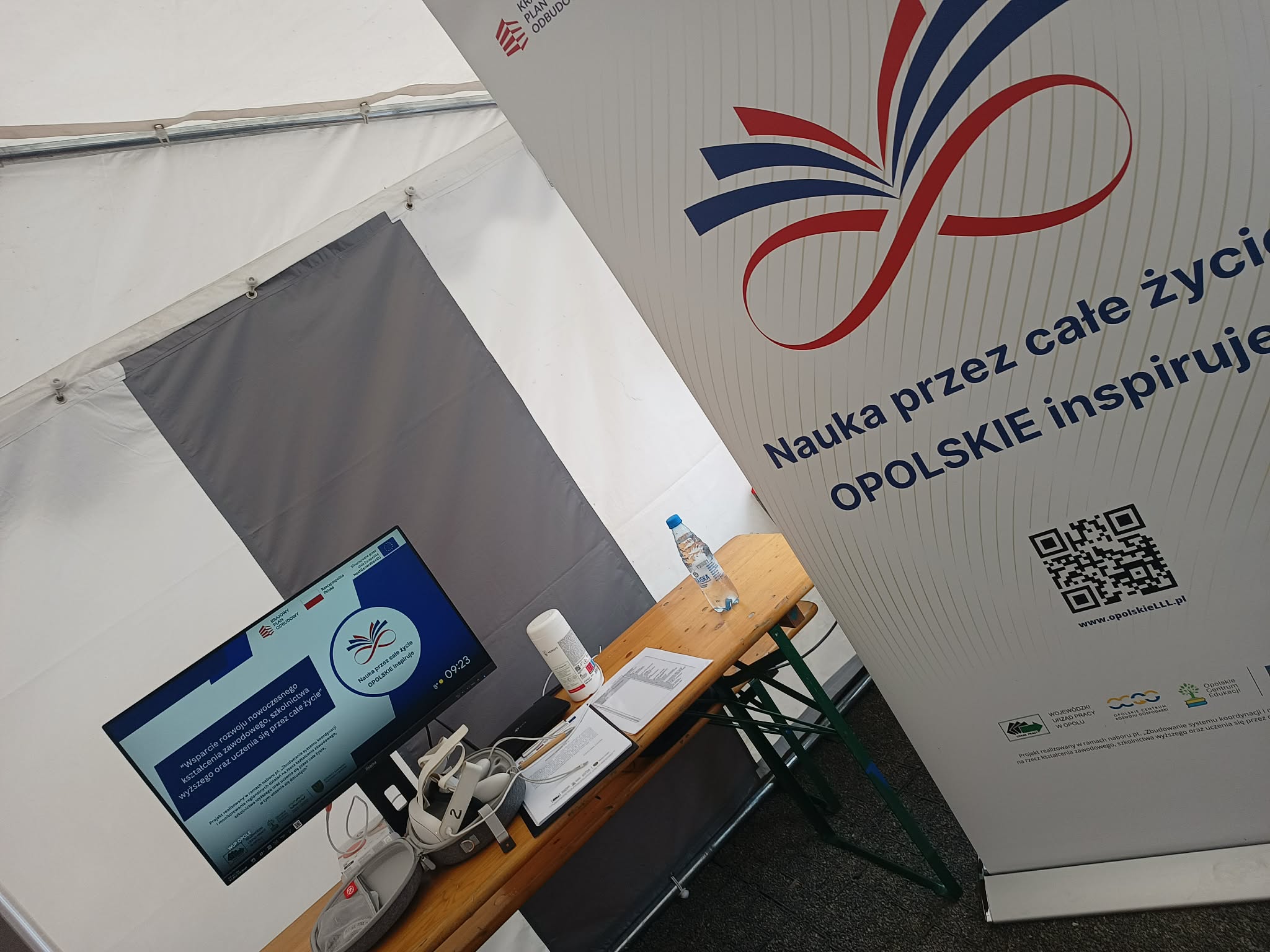 Stół wewnątrz namiotu mieści monitor komputerowy, zestaw VR, butelkę wody i inne przedmioty. Obok stoi wysoki baner z czerwono-niebieskim symbolem nieskończoności i polskim tekstem promującym uczenie się przez całe życie na Opolszczyźnie.