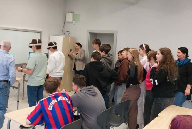 Grupa młodzieży szkolnej oraz dorosłych obserwuje dwóch uczniów korzystających z gogli VR w jasnej klasie. Uczniowie z goglami stoją przy stanowiskach z monitorami, a reszta grupy przygląda się ich działaniom z zainteresowaniem. W tle widoczny jest interaktywny ekran oraz biała tablica, a jeden z uczniów na pierwszym planie ma na sobie koszulkę piłkarską Bayernu Monachium.