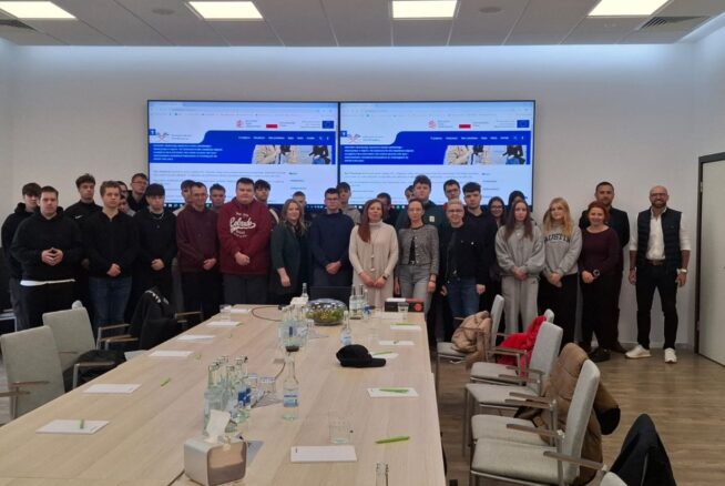 Grupa około 22 osób, głównie młodych dorosłych, stoi razem w sali konferencyjnej. Przed nimi stoją stoły konferencyjne z butelkowaną wodą i notatnikami. Dwa duże ekrany wyświetlają stronę internetową z niebieskimi i białymi akcentami.