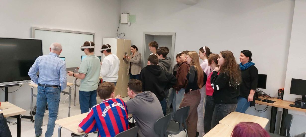 Grupa młodzieży szkolnej oraz dorosłych obserwuje dwóch uczniów korzystających z gogli VR w jasnej klasie. Uczniowie z goglami stoją przy stanowiskach z monitorami, a reszta grupy przygląda się ich działaniom z zainteresowaniem. W tle widoczny jest interaktywny ekran oraz biała tablica, a jeden z uczniów na pierwszym planie ma na sobie koszulkę piłkarską Bayernu Monachium.