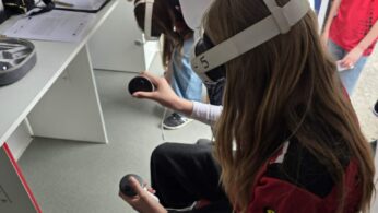 Zbliżenie na osobę siedzącą wewnątrz mobilnego laboratorium, która ma na sobie białe gogle VR i trzyma w dłoniach bezprzewodowe kontrolery. W tle widoczne jest stanowisko z monitorem oraz fragmenty dokumentacji leżącej na białym blacie. Obok stoi inna osoba pomagająca w obsłudze sprzętu. Całość prezentuje praktyczne wykorzystanie technologii wirtualnej rzeczywistości.
