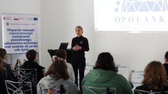 Grupa nastolatków siedzi na metalowych krzesłach w jasnej sali konferencyjnej o nowoczesnym designie. Po lewej stronie widać rząd krzeseł z zawieszonymi granatowymi torbami z białym logo „OPOLANKA”. Po prawej stronie kadru, przy wysokim czarnym stoliku, stoi kobieta w ciemnym stroju, która prowadzi prezentację. Obok niej znajduje się rollup z logotypami i tekstem dotyczącym wspierania kształcenia zawodowego w powiecie nyskim.