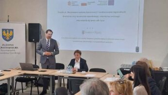 Zdjęcie przedstawia spotkanie w jasnej sali konferencyjnej, gdzie mężczyzna w szarym garniturze stoi i przemawia przy ekranie projekcyjnym. Na ekranie widoczny jest tytuł projektu dotyczącego budowy systemu koordynacji kształcenia zawodowego w województwie opolskim. Uczestnicy siedzą przy stołach ustawionych w podkowę, słuchając prezentacji i śledząc treści na swoich laptopach.