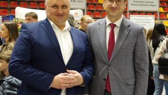 Dwóch uśmiechniętych mężczyzn w garniturach stoi blisko siebie na pierwszym planie w zatłoczonej hali. Mężczyzna po lewej ma na sobie granatową marynarkę, białą koszulę i splecione dłonie. Mężczyzna po prawej nosi szary garnitur w kratę, czerwoną koszulę i okulary. W tle widać rozmyte sylwetki innych ludzi i trybuny hali sportowej.