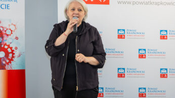 Kobieta z krótkimi siwymi włosami, ubrana w czarną kurtkę, stoi i mówi do mikrofonu. Za nią znajdują się banery z czerwonym i niebieskim logo i tekstem, wskazujące na oficjalne lub profesjonalne otoczenie wydarzenia.