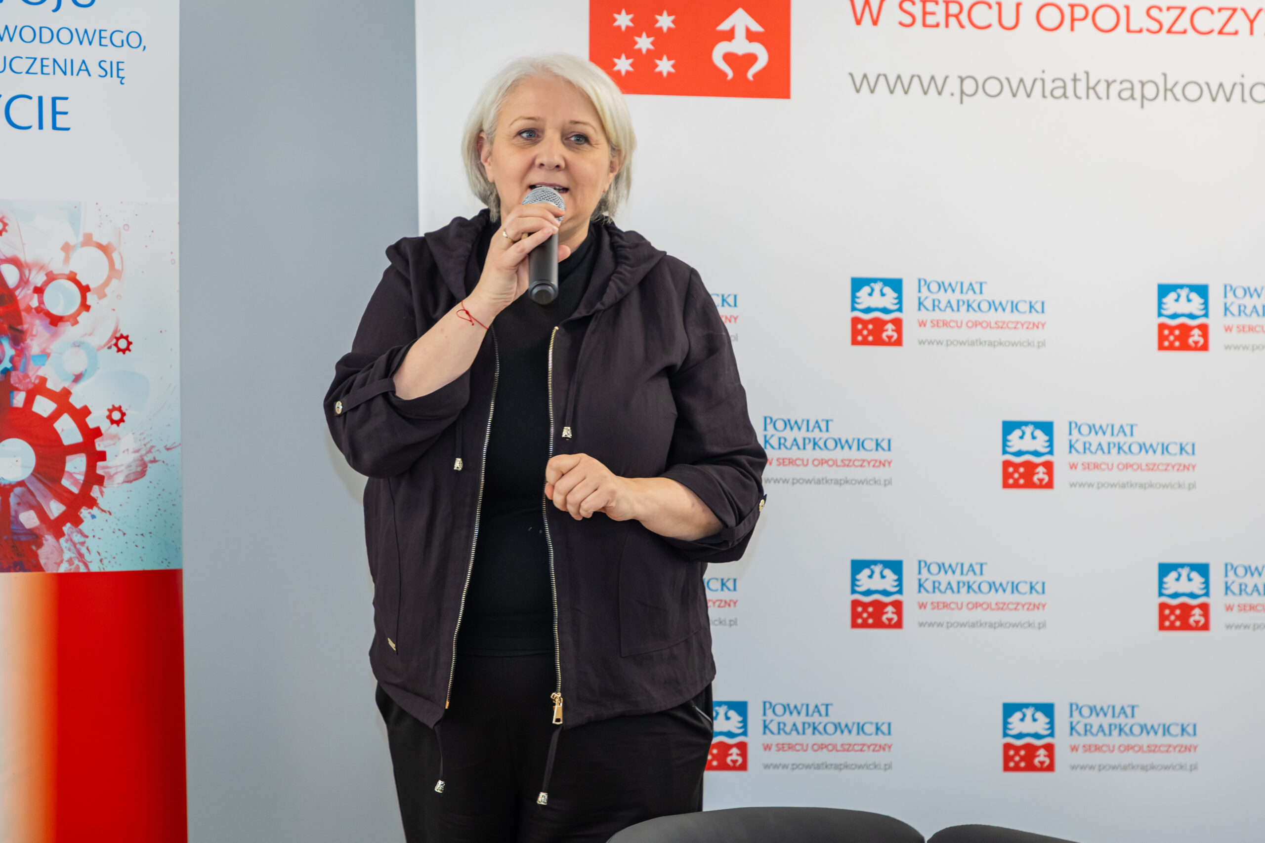 Kobieta z krótkimi siwymi włosami, ubrana w czarną kurtkę, stoi i mówi do mikrofonu. Za nią znajdują się banery z czerwonym i niebieskim logo i tekstem, wskazujące na oficjalne lub profesjonalne otoczenie wydarzenia.