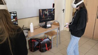 Dwoje młodych ludzi w klasie nosi białe zestawy VR. Jeden z nich stoi w pobliżu monitora i biurka z butelką wody i torbami pod spodem. W tle widoczna jest tablica, szafki i zamknięte drzwi. Oboje są zaangażowani w wirtualne doświadczenie.