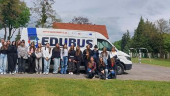 Grupa młodzieży stoi w rzędzie przed białym furgonetką z dużym napisem „EDUBUS” oraz adresem strony internetowej. W tle widać zielone drzewa, fragment budynku z czerwonym dachem oraz metalową konstrukcję bramki na asfalcie. Część osób stoi bezpośrednio przed pojazdem, a trzy dziewczyny kucają na pierwszym planie po prawej stronie. Wszyscy są ubrani w codzienne, wiosenne stroje, a cała scena rozgrywa się na zewnątrz w pochmurny dzień.