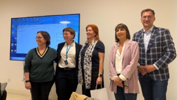 Grupa pięciu uśmiechniętych osób w eleganckich strojach biznesowych stoi obok siebie w jasnym pomieszczeniu. W tle znajduje się duży monitor wyświetlający niebieski ekran systemowy z tekstem i ikonami. Jedna z kobiet ma na szyi niebieską smycz z identyfikatorem.