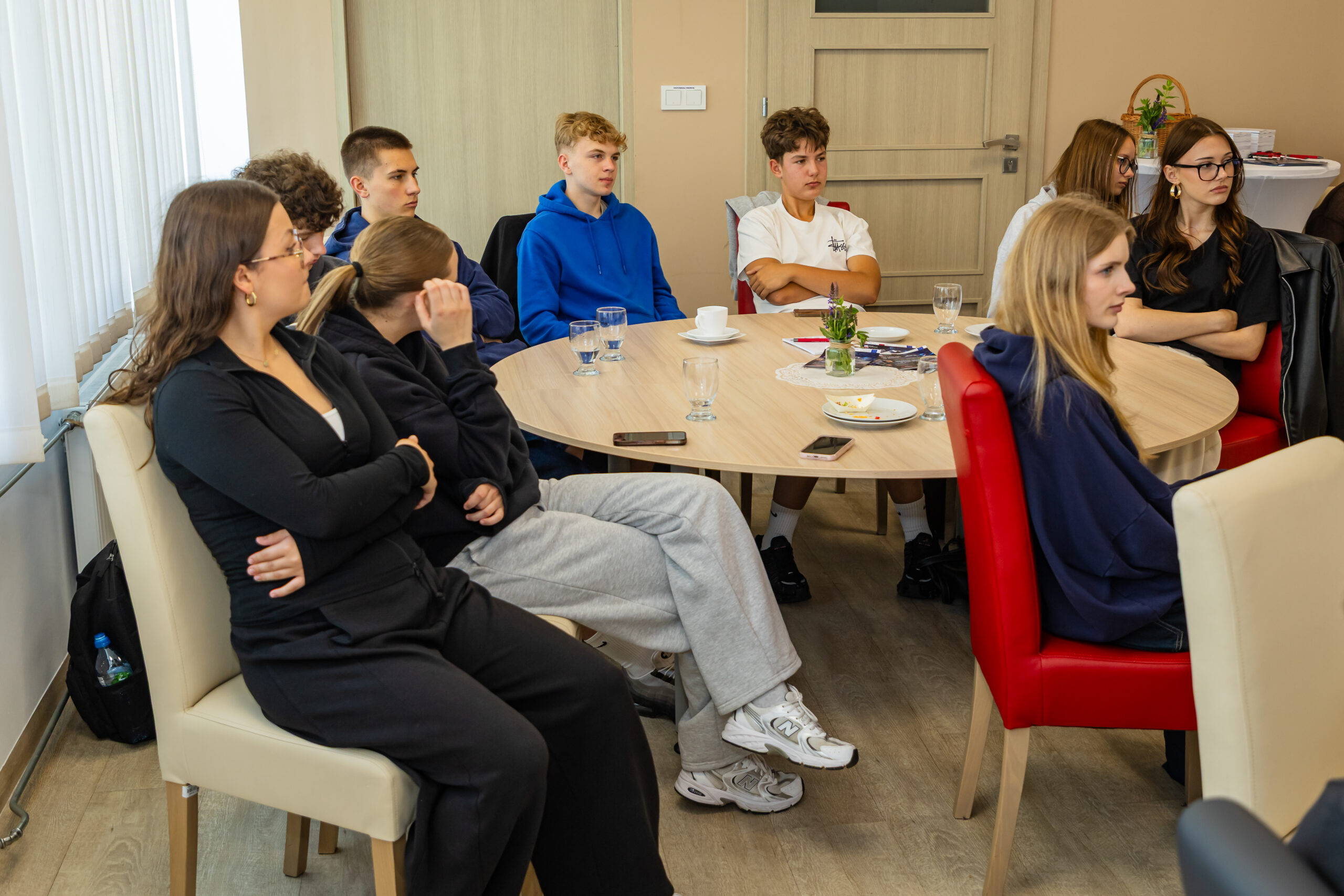 Grupa młodych ludzi siedzi wokół okrągłego stołu w swobodnej sali konferencyjnej i uważnie słucha. Większość z nich ma na sobie zwykłe ubrania, takie jak bluzy z kapturem i spodnie dresowe. Na stole leżą puste szklanki, talerze i papiery. Nastrój wydaje się skupiony i zaangażowany.