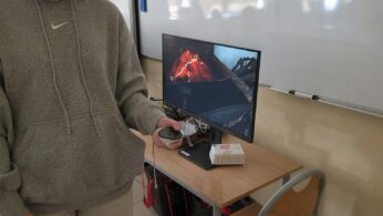 Osoba w szarej bluzie z kapturem i spodniach dresowych używa białego zestawu VR i kontrolera w klasie. Za nimi monitor na biurku wyświetla ognistą scenę wulkanu. Na biurku znajduje się klawiatura, mysz, zestaw słuchawkowy, a obok tablica i puste krzesło.
