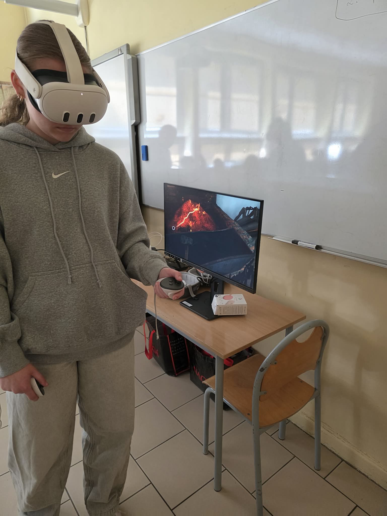 Osoba w szarej bluzie z kapturem i spodniach dresowych używa białego zestawu VR i kontrolera w klasie. Za nimi monitor na biurku wyświetla ognistą scenę wulkanu. Na biurku znajduje się klawiatura, mysz, zestaw słuchawkowy, a obok tablica i puste krzesło.