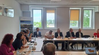 Grupa ludzi, głównie w strojach biznesowych, siedzi wokół prostokątnych stołów w jasnej sali konferencyjnej z trzema dużymi oknami. Niektórzy rozmawiają, inni słuchają. Na stołach leżą laptopy, papiery i kubki. Widoczne są dwa stojące banery i znak na ścianie.