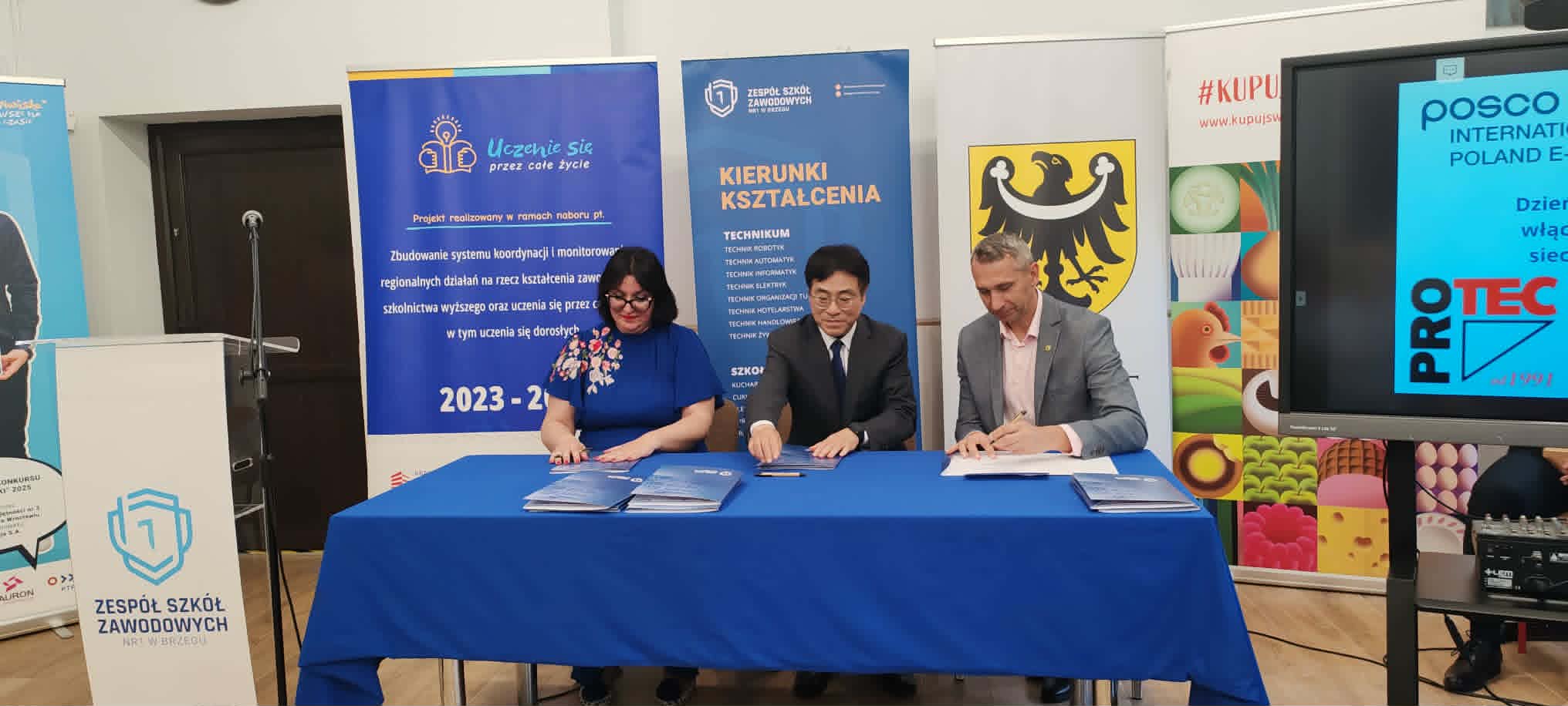 Trzy osoby siedzą przy stole podpisując dokumenty na oficjalnym wydarzeniu. Za nimi znajdują się banery z polskim tekstem, logo szkoły i regionu oraz logo firmy ProTec. Otoczenie wydaje się formalne, prawdopodobnie w szkole lub instytucji edukacyjnej.